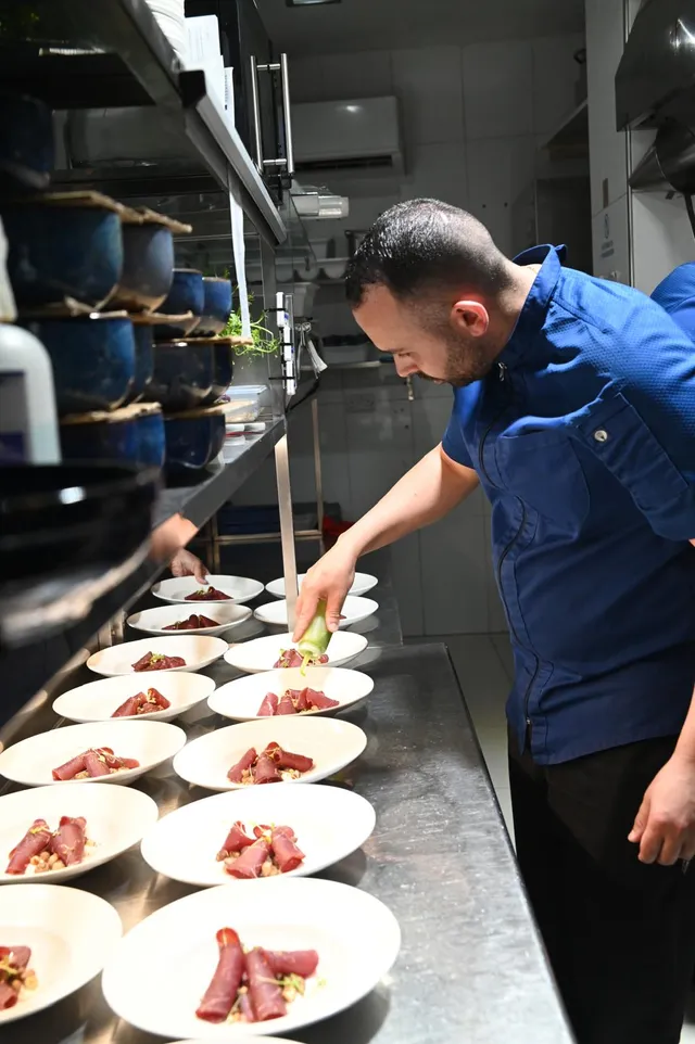 A chef in the kitchen dressing a long line of plated tuna rolls, each finished with micro herbs
