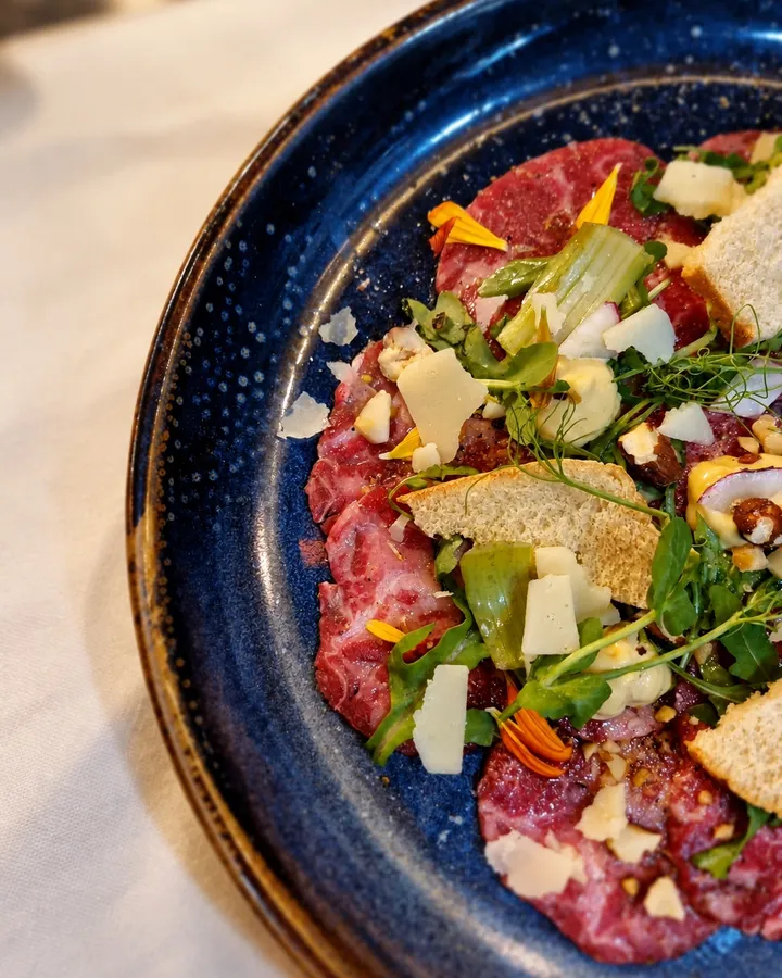 Beef carpaccio on a blue speckled plate with rocket, shaved cheese, croutons and orange petals