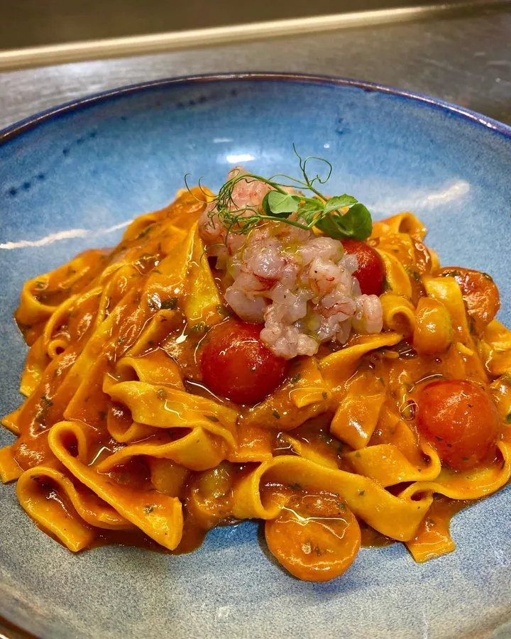 The same tagliatelle seen closer: prawn tartare and pea shoots over pasta in tomato sauce, in a blue bowl