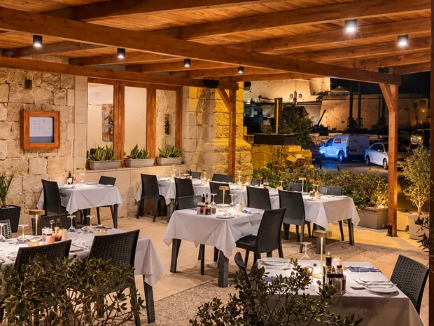 Acqua Blu's terrace after dark: tables laid with white linen under a timber pergola, lamps lit, olive trees in planters along its edge