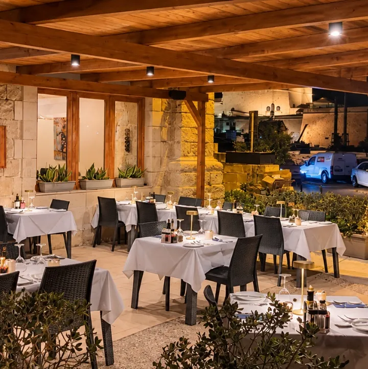 Acqua Blu's terrace after dark: tables laid with white linen under a timber pergola, lamps lit, olive trees in planters along its edge